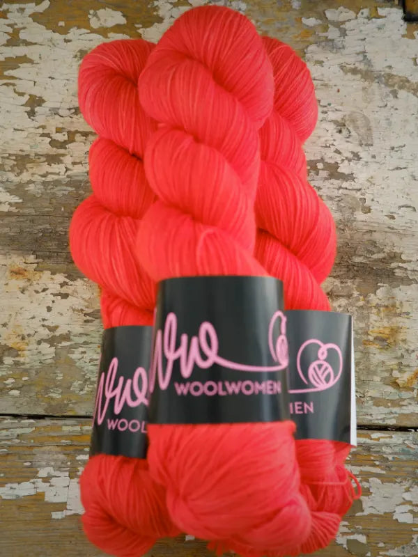 Two skeins of bright coral hand-dyed WoolWomen Merino Sock with black paper labels.