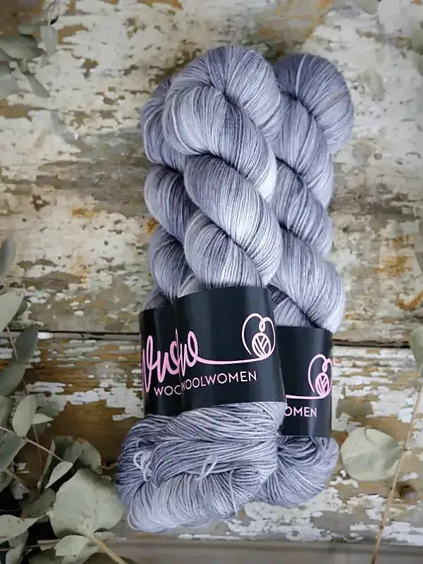 Two skeins of silver-grey WoolWomen Merino Sock yarn with a subtle heathered texture, wrapped in black labels.