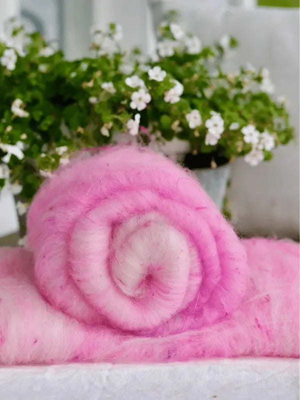A fluffy, rolled-up WoolWomen Art Batt - Corriedale in pink rests on matching folded wool. In the background, a blurred potted plant with small white flowers and green leaves adds a natural touch.