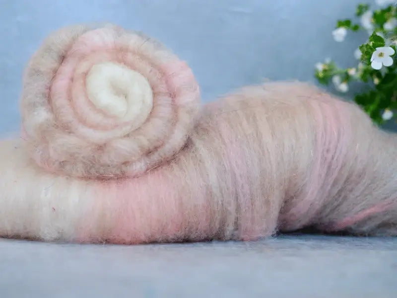 A soft, fluffy bundle of käsinvärjätty villa and sari silkki in pale pink and cream, rolled into a spiral, rests on a light surface with white flowers and green leaves. Featured: WoolWomen Art Batt -BFL.