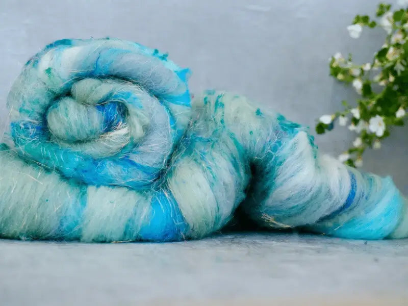A close-up of a rolled WoolWomen Art Batt -Cormo in blue and teal, showcasing its soft, fluffy texture. Small white flowers and green leaves appear blurred in the right-side background.