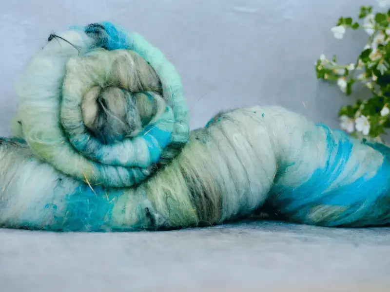 A rolled bundle of WoolWomen Art Batt -Cormo in blue, green, and white rests on a gray surface with white flowers behind—ideal as an art batt or kehräyskuitu for creative projects.