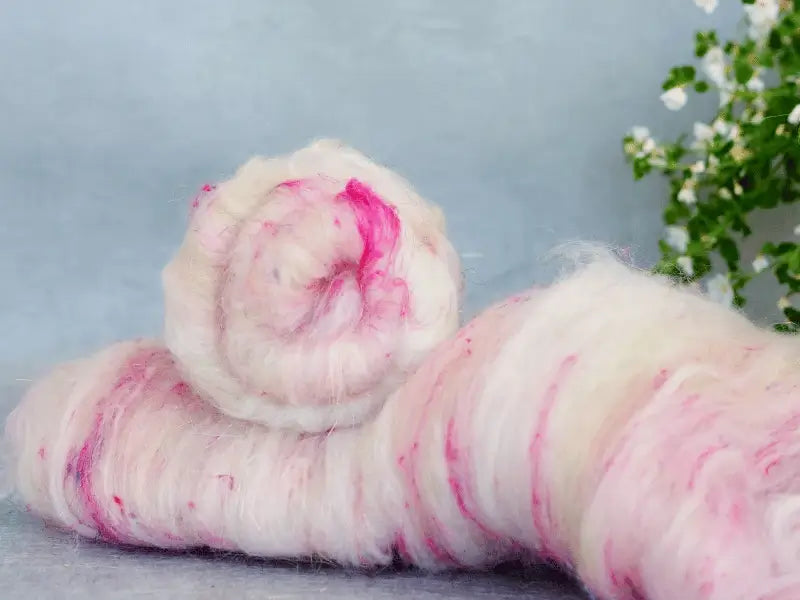 A close-up of soft, fluffy WoolWomen Art Batt -Polwarth in white with pink streaks, rolled on a surface with a small green plant and white flowers in the background—perfect for spinning.