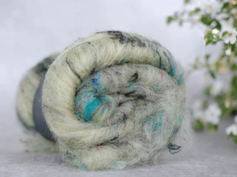 A close-up of a rolled bundle of WoolWomen Art Batt - Suomalainen villa, soft and fuzzy in light grey with blue and black hints, resting on a light surface with small white flowers softly blurred in the background.