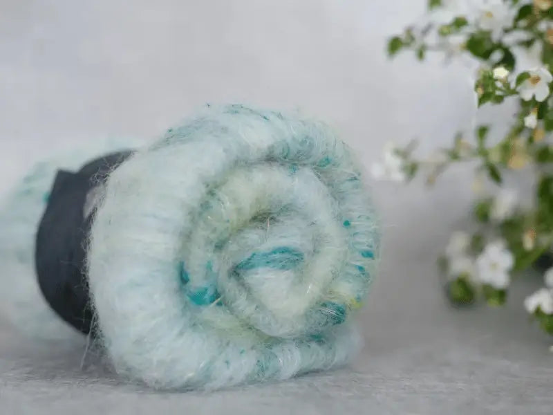 A close-up of a rolled, soft, pastel blue WoolWomen Art Batt - Suomalainen villa fiber with green and teal tones on a gray surface. Blurred white flowers with green leaves appear in the background on the right.