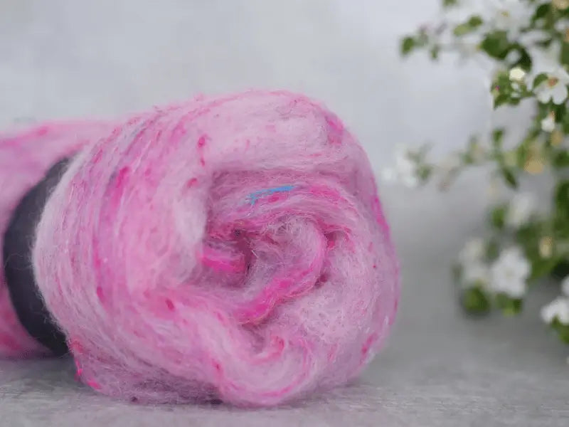 A close-up of a roll of WoolWomen Art Batt - Suomalainen villa in soft pink and purple with colorful flecks, resting on a gray surface. White flowers and green leaves are softly blurred in the background to the right.