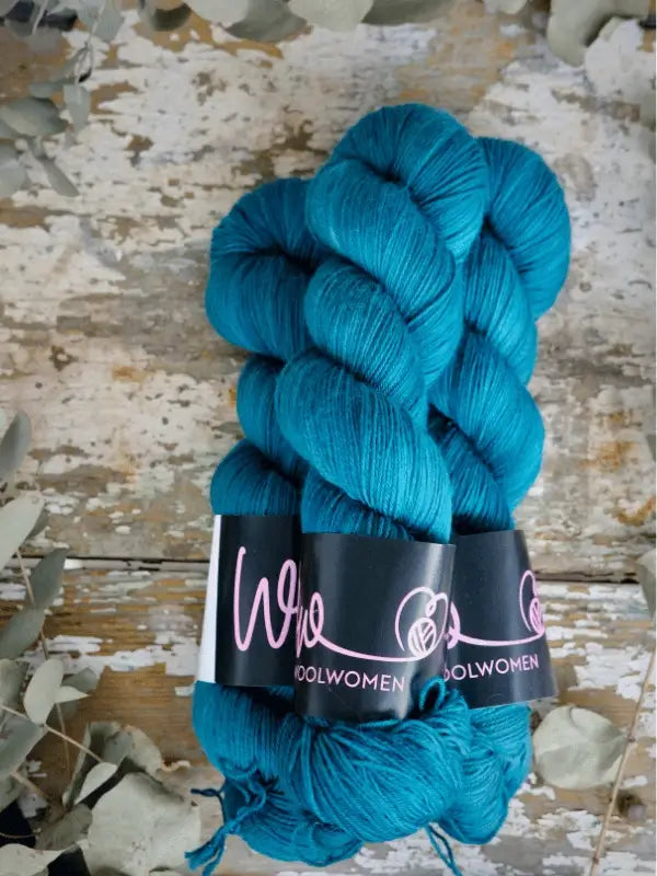 Two skeins of bright teal WoolWomen Merino Sock, each with a black WOOLWOMEN label, rest on a rustic wooden surface surrounded by dried leaves.