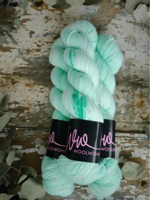 Two skeins of pale mint-green WoolWomen Merino Sock yarn with subtle darker green streaks, wrapped in black "WoolWomen" bands, rest on a rustic wooden surface scattered with dried leaves. Soft superwash merinovilla.