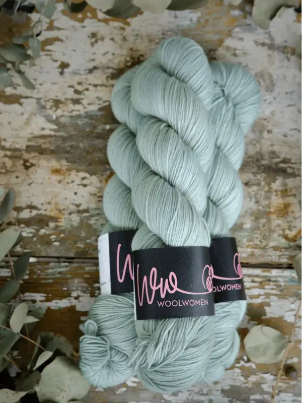 Two skeins of WoolWomen Merino Sock käsinvärjätty pale blue-green yarn sit on a distressed wooden surface beside dried eucalyptus leaves.
