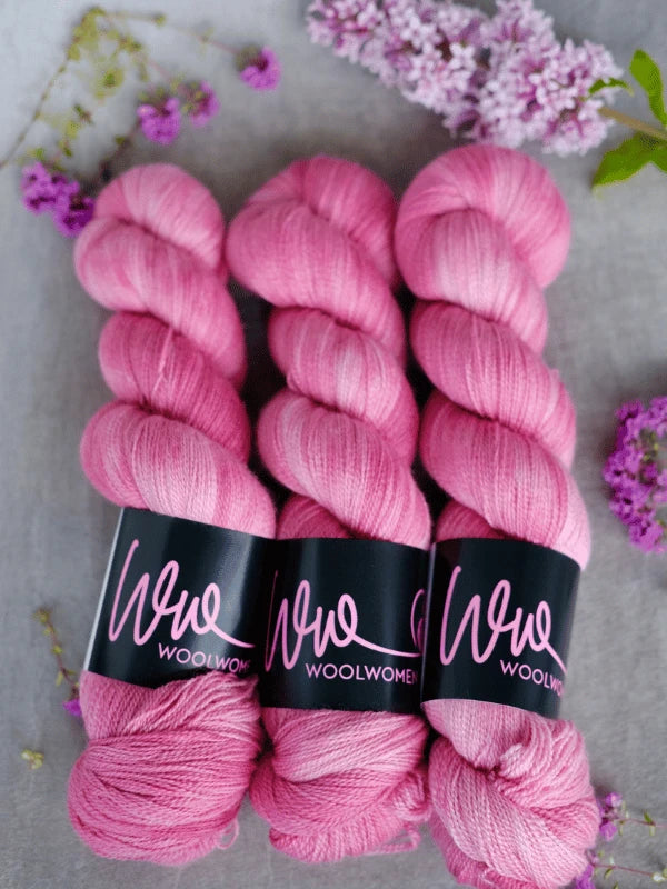 Three skeins of pink WoolWomen Merino Silk Lace yarn with black “Wool Women” labels are lined up on a light surface, surrounded by small purple flowers and lilac blossoms.