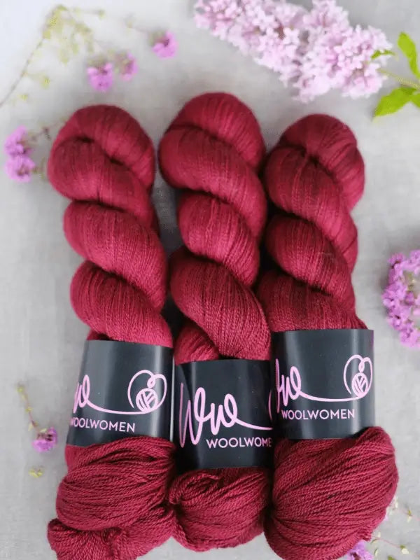 Three skeins of rich red WoolWomen Merino Silk Lace are arranged side by side on a light surface, surrounded by small pink and purple flowers and lilac blossoms.