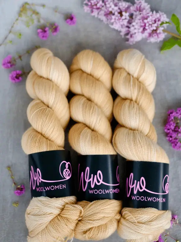 Three skeins of pale yellow WoolWomen Merino Silk Lace yarn with Woolwomen labels are arranged side by side on a light surface, surrounded by small purple and lilac flowers.