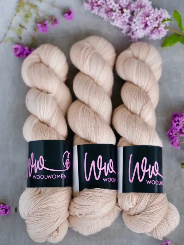 Three skeins of pale beige WoolWomen Merino Silk Lace yarn lie side by side on a gray surface, surrounded by small purple flowers and sprigs of lilac.
