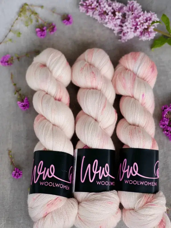 Three skeins of WoolWomen Merino Silk Lace in pale pink are arranged side by side on a gray surface, surrounded by purple flowers and lilac blossoms.