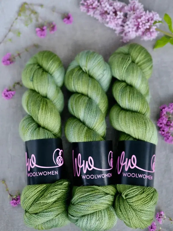 Three skeins of green WoolWomen Merino Silk Lace yarn, including käsinvärjätty merinovilla, are arranged side by side on a gray surface with small purple flowers and lilac blossoms scattered around.