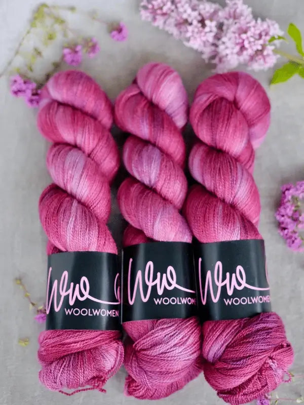 Three skeins of WoolWomen Merino Silk Lace in pink and purple variegated tones are placed side by side on a light surface, accented with small purple flowers scattered around them.