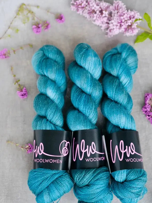 Three skeins of teal blue WoolWomen Merino Silk Lace yarn are arranged on a light surface, surrounded by small lilac and pink flowers and petals.