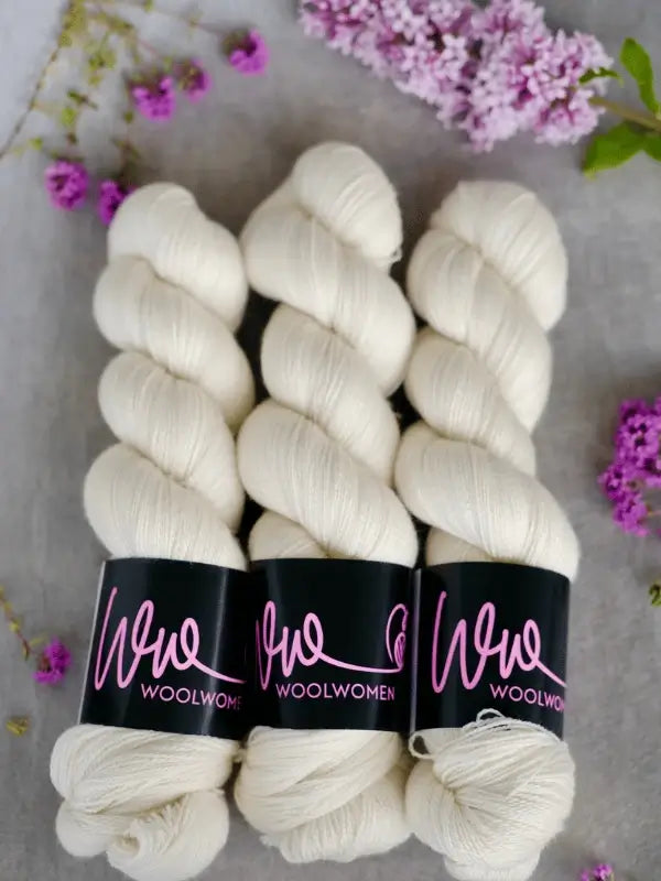 Three skeins of WoolWomen Merino Silk Lace yarn are arranged side by side on a soft surface, surrounded by small purple and lilac flowers.