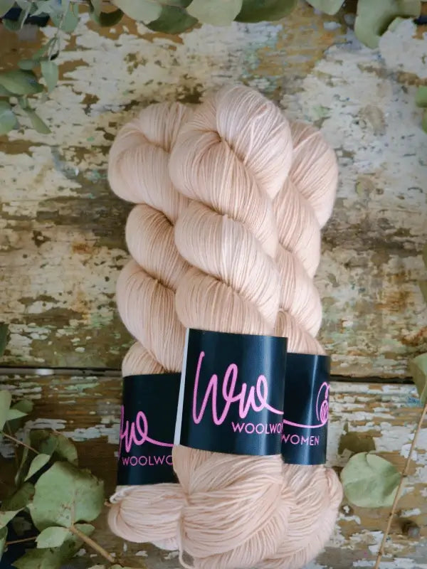 Two skeins of pale beige WoolWomen Merino Sock yarn, made from superwash merino wool, rest on a distressed wooden surface surrounded by scattered green leaves.
