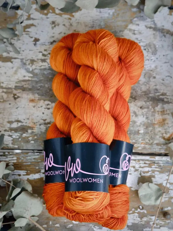 Three skeins of WoolWomen Merino Sock käsinvärjätty orange yarn with black labels are arranged on a rustic wooden surface, surrounded by scattered dried leaves.