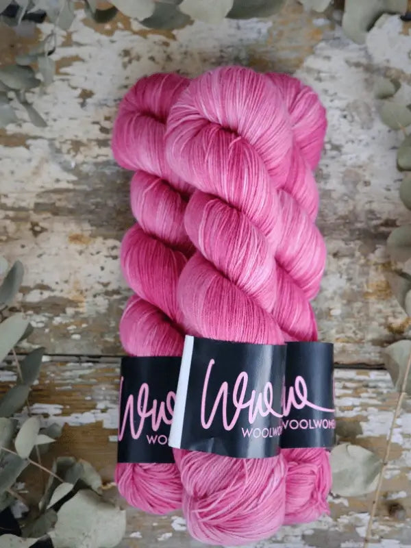 Two skeins of pink WoolWomen Merino Sock yarn with black and white WoolWoolWoolWong labels lie side by side on a rustic wooden surface, surrounded by greenish-gray leaves.