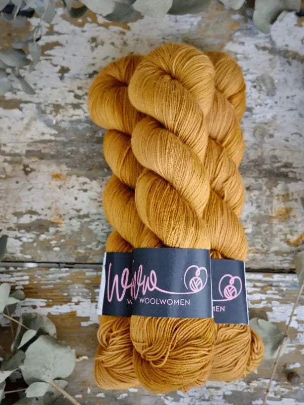 Two skeins of golden brown WoolWomen Merino Sock yarn with black labels rest on a weathered wooden surface, surrounded by scattered dried leaves.