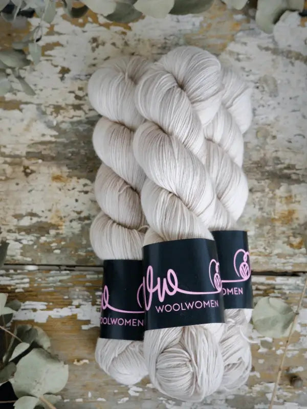 Three skeins of soft, off-white WoolWomen Merino Sock yarn with pink script labels rest on a rustic wooden surface, surrounded by pale green eucalyptus leaves.
