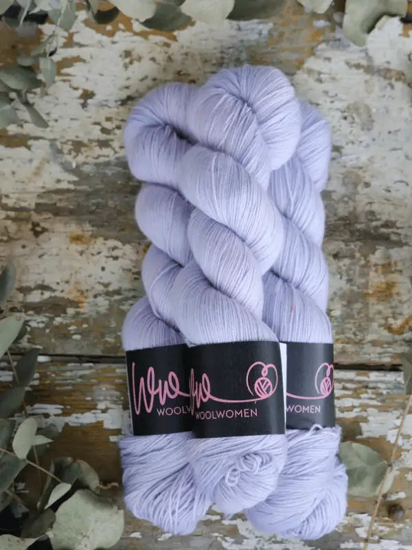 Three skeins of WoolWomen Merino Sock yarn in light lavender, featuring black labels, are displayed on a rustic wooden surface surrounded by eucalyptus leaves.
