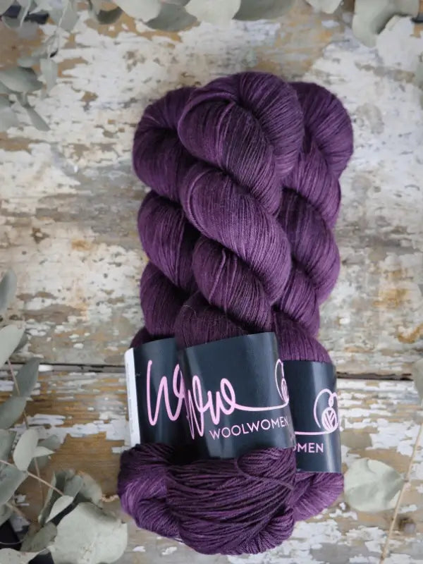 Two skeins of deep purple WoolWomen Merino Sock yarn with black labels rest on a rustic wooden surface amid dried leaves. These superwash merinovilla yarns bring richness and elegance to your next project.