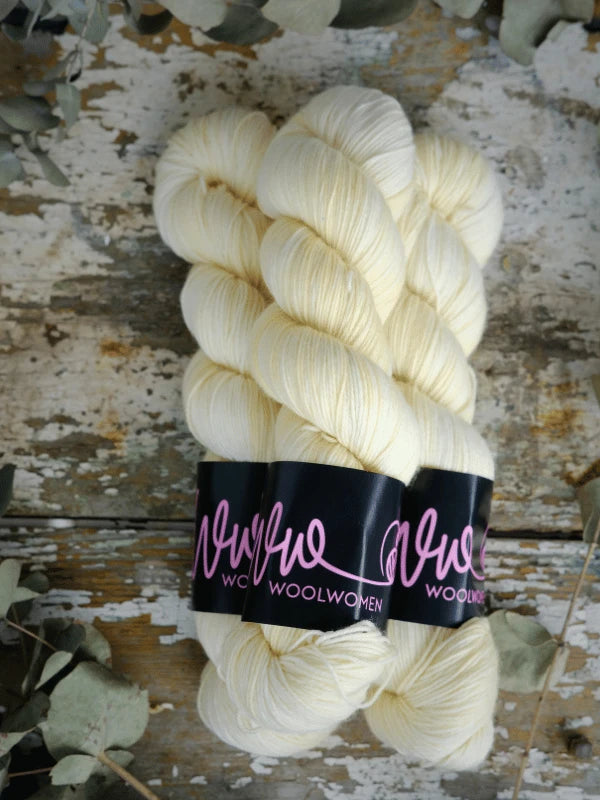 Two skeins of WoolWomen Merino Sock, soft off-white superwash merinovilla yarn with black Wool Women labels, displayed on a rustic wooden surface surrounded by dried leaves.