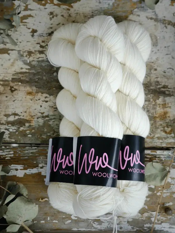 Two skeins of soft, white WoolWomen Merino Sock yarn with black labels in pink script rest on a rustic wooden surface, surrounded by scattered dried leaves.