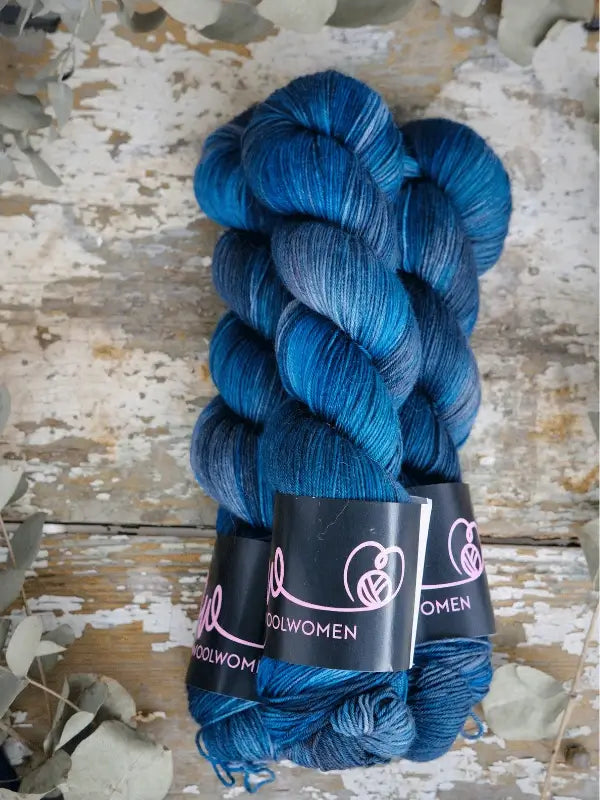 Three skeins of WoolWomen Merino Sock in deep blue, each wrapped with black labels featuring pink branding, are displayed on a rustic wooden surface with dried leaves at the edges.