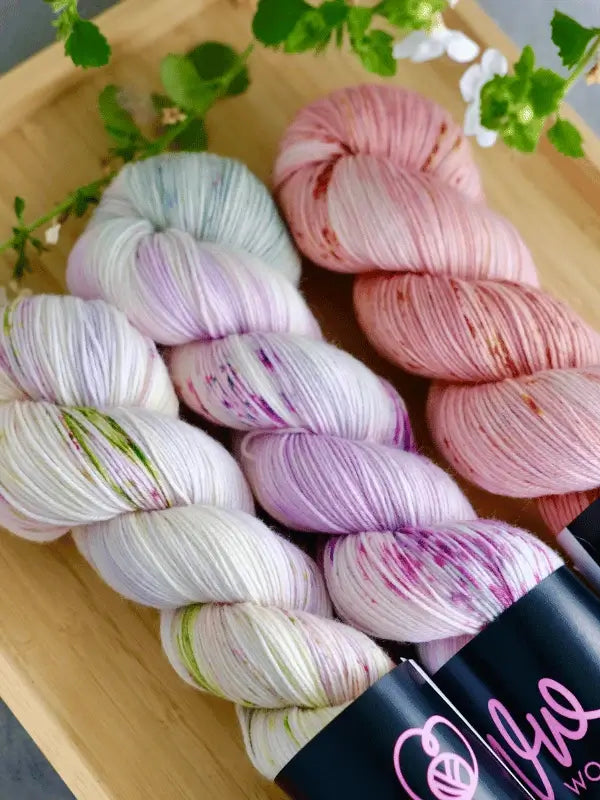 Three skeins of WoolWomen merino sock - Summer Collection yarn in pastel pink, purple, green, and white are arranged on a wooden tray with green leaves and small white flowers.