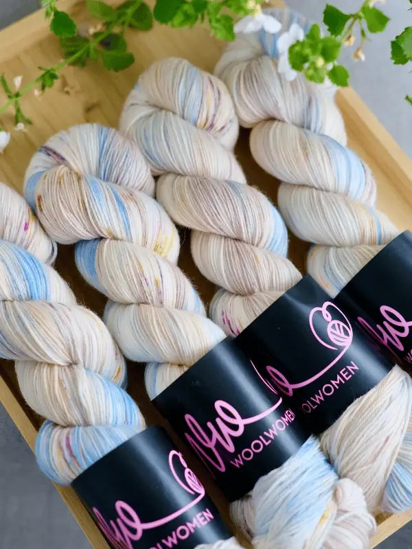Four skeins of WoolWomen merino sock - Summer Collection yarn with pastel blue and beige tones, each wrapped in a black "WoolWomen" label, arranged neatly in a wooden tray beside green leaves.