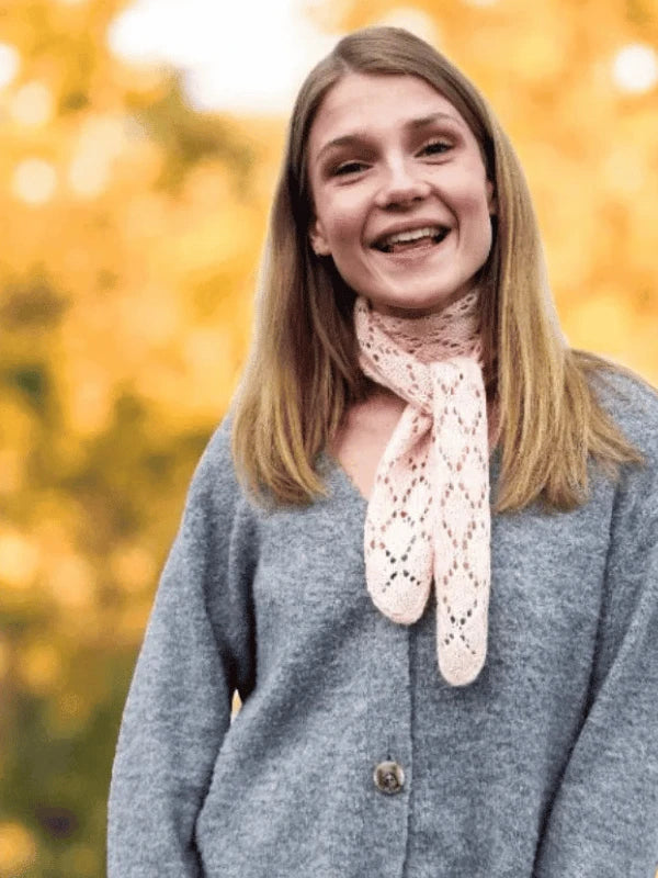 A young woman with straight blonde hair smiles outdoors, wearing a gray cardigan and a light pink knit scarf with an I-cord reunus. Blurred golden autumn foliage completes the scene—perfect inspiration for your next WoolWomen MinMax huiviohje!.