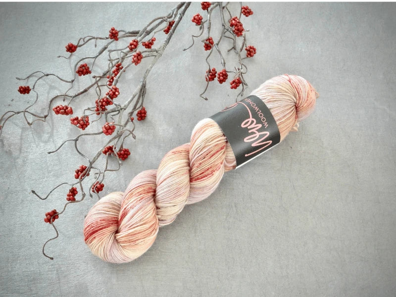 A skein of WoolWomen Merino Sock – Yhden Padan Ihme, a käsinvärjätty mulesing-vapaa merino yarn in light pink and cream with red speckles, rests on gray next to a branch of small red berries—a perfect choice for your next soft creation.