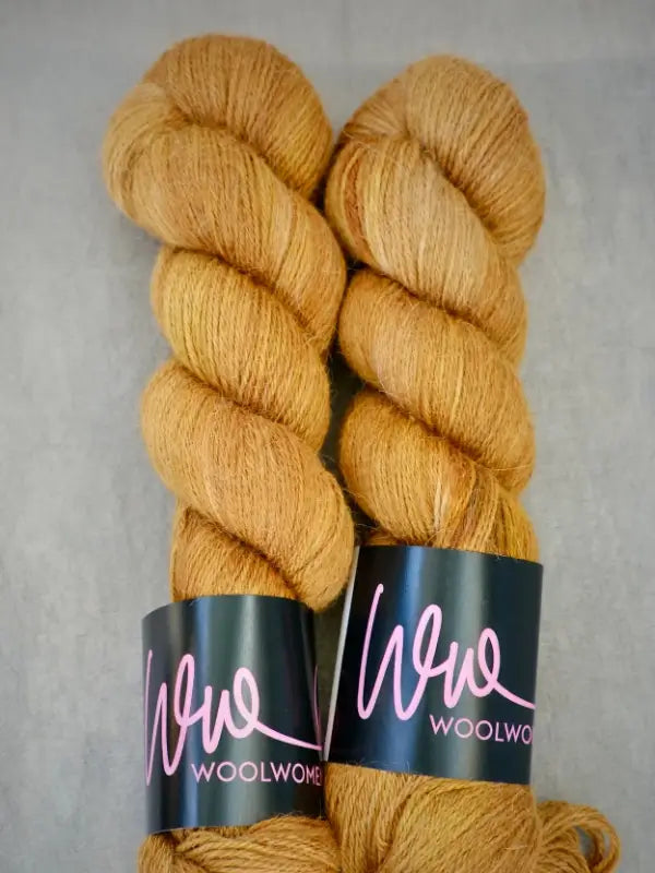 Two skeins of golden-brown yarn lie side by side on a gray surface. Each skein features a black label adorned with “WoolWomen AlpaLux” in pink cursive lettering, highlighting the luxurious quality of this ylellinen lanka crafted from soft baby alpakka.