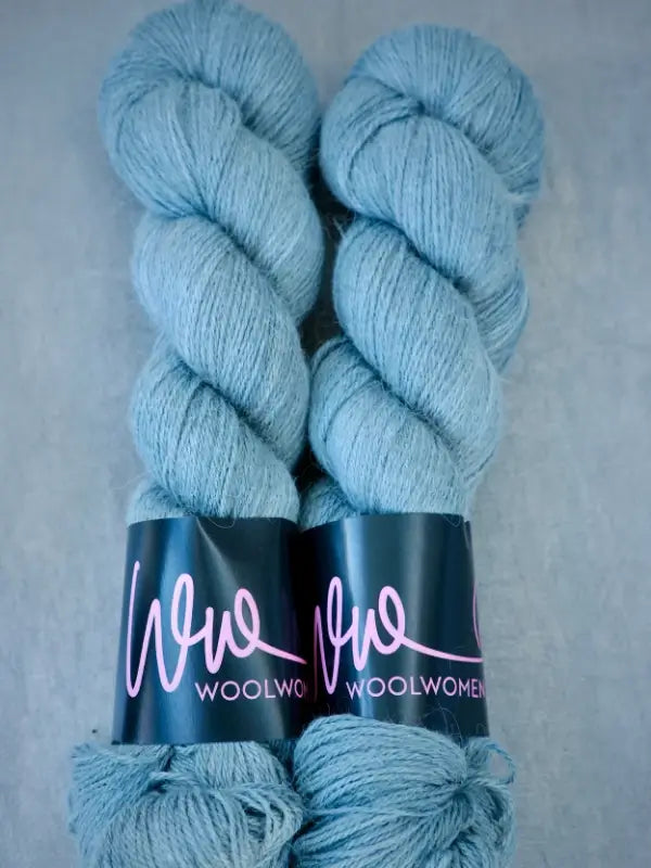 Two skeins of light blue WoolWomen AlpaLux alpakka-lanka yarn with black labels featuring pink “WoolWomen” lettering, lying side by side on a gray surface.