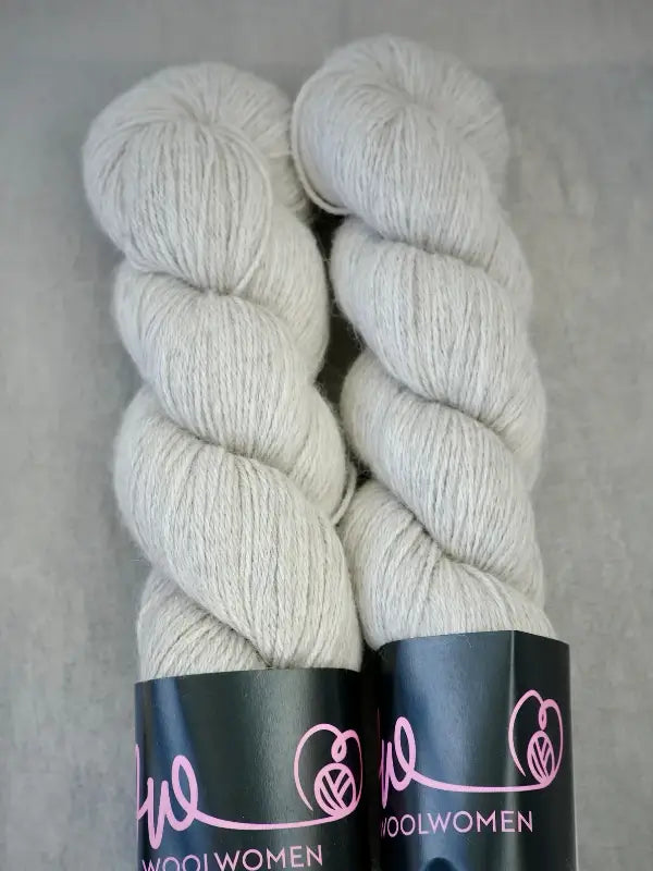 Two skeins of soft, pale gray WoolWomen AlpaLux baby alpakka yarn with WOOLWOMEN branding on black labels, placed side by side on a neutral background.