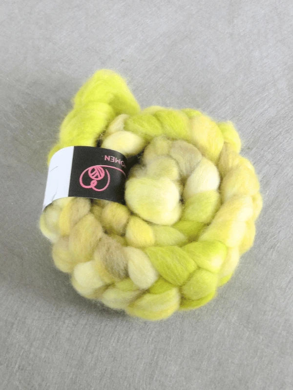 A coiled bundle of thick, soft, yellow and light green WoolWomen Suomalainen Kehruuvilla käsinvärjätty wool roving rests on a light gray surface, wrapped with a black label featuring white and pink text.
