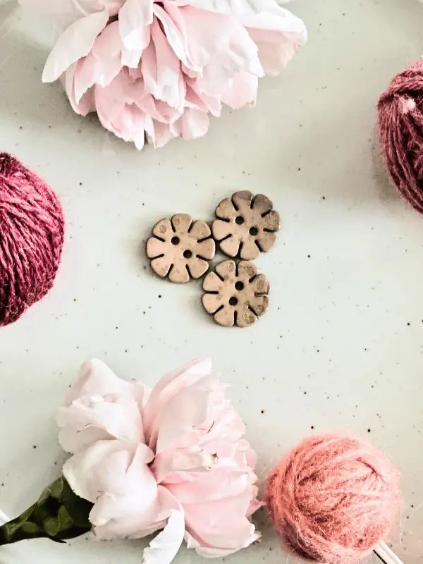 Three Värikkäät kukkanapit 20mm (17700) are displayed on a white surface, accented by two pink peonies and two balls of pink yarn for a yksinkertainen, elegant look.