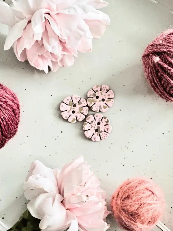 Two pink peonies, two balls of pink yarn, and four Värikkäät kukkanapit 20mm (17700) are arranged on a light gray speckled surface for a simple yet charming display.