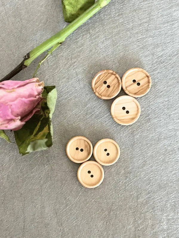 Four pairs of Puunappi laakea 18mm ja 20mm (7930), light wooden buttons with two holes each, are arranged on a gray textured surface beside a dried pink rose.