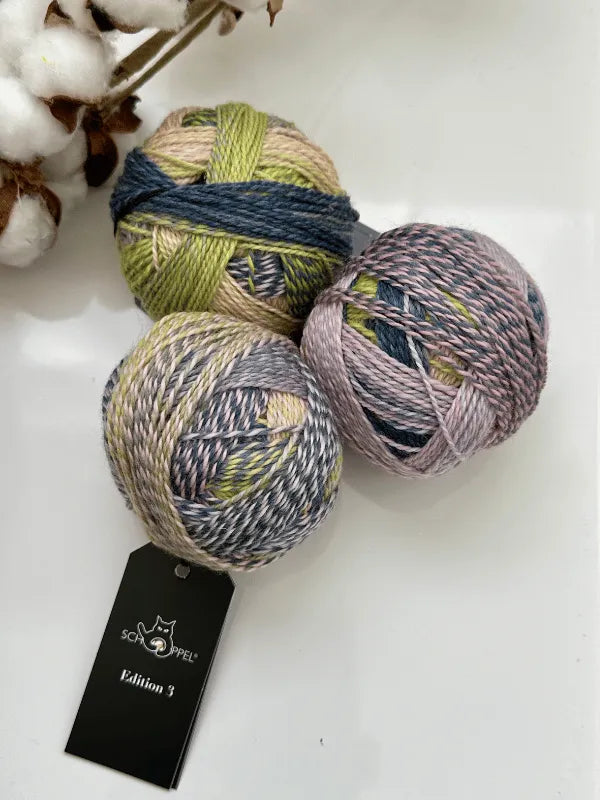 Three multicolored balls of Schoppel-Wolle Edition 3 yarn in green, blue, and gray rest on a white surface beside a cotton plant. One ball with a black tag is made from superwash merino wool.