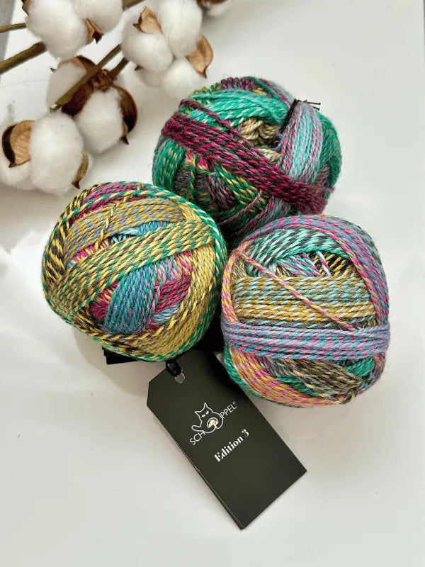 Three colorful balls of Schoppel-Wolle Edition 3 superwash-merino yarn are displayed on a white surface near a cotton branch. One ball features a black label with a cat logo and the text "Schoppel-Wolle Edition 3.