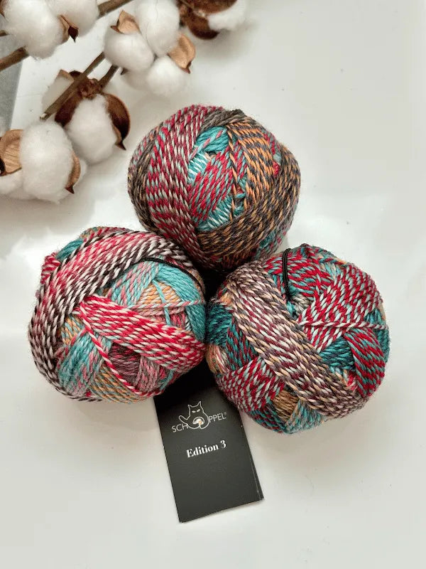 Three multicolored balls of Schoppel-Wolle Edition 3 yarn rest on a white surface beside a black label, with cotton branches displaying fluffy white bolls in the background.