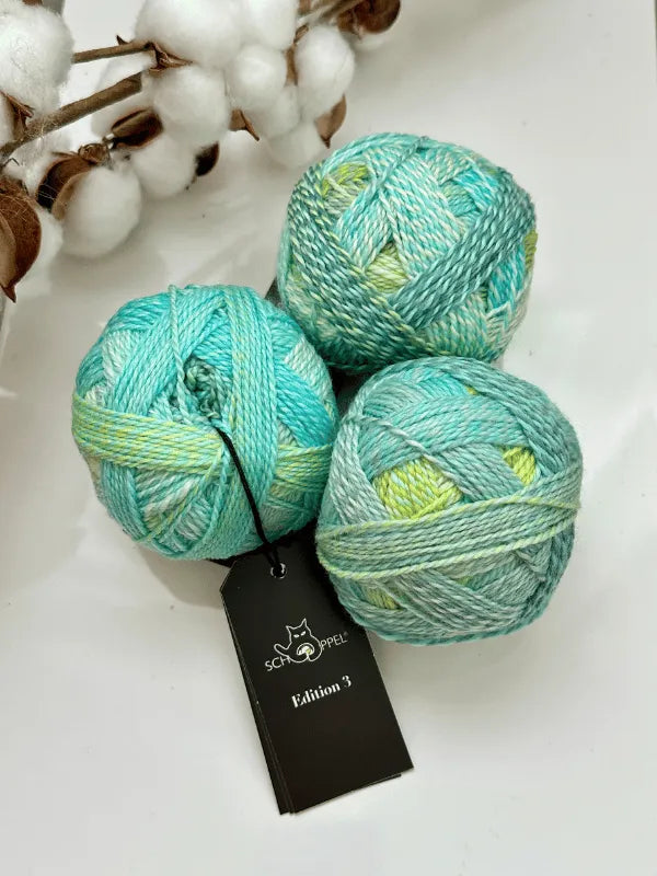Three balls of Schoppel-Wolle Edition 3 yarn in turquoise, green, and cream rest on a white surface beside cotton stems. One soft superwash merino wool ball has a black Schoppel-Wolle Edition 3 tag attached.