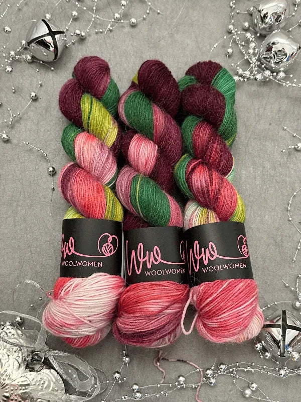 Three skeins of WoolWomen Alpaca Sock - yhden padan ihmeet yarn in green, burgundy, pink, and yellow are neatly arranged on a gray surface with silver bells and sparkly string. Ideal for lämmin sukalanka projects.
