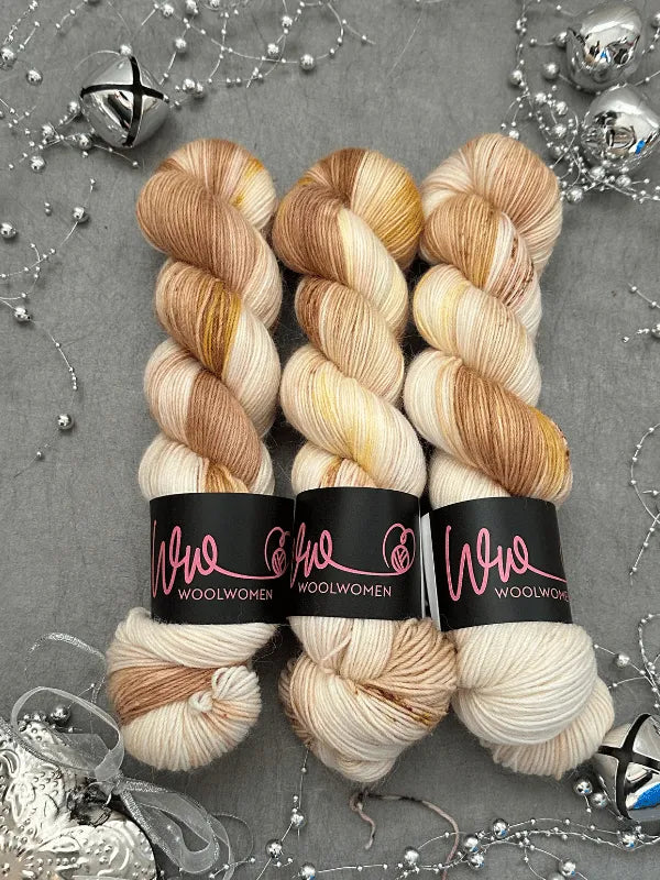 Three skeins of cream, beige, and light brown yarn—ideal for making WoolWomen Alpaca Sock -yhden padan ihmeet—are displayed side by side on a gray surface adorned with silver string, bells, and festive ornaments.