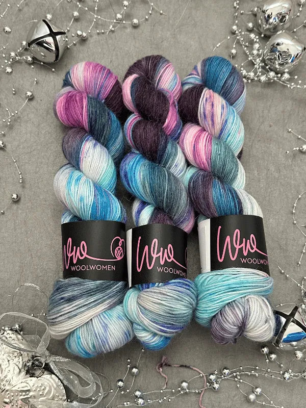 Three skeins of WoolWomen Alpaca Sock -yhden padan ihmeet yarn in blue, purple, pink, and white are arranged on a gray surface adorned with silver beads and ornaments.